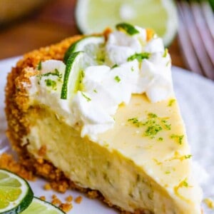 A slice of key lime pie with a graham cracker crust, topped with whipped cream, lime zest, and lime slices, sits on a white plate with extra lime slices beside it.
