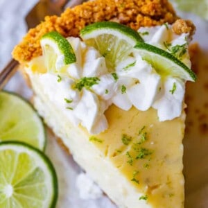 A close-up of a key lime pie slice on a white plate, topped with whipped cream and lime slices, featuring a graham cracker crust with extra lime on the side.