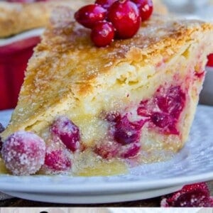 A slice of cranberry pie with a creamy custard filling and sugared cranberries on top sits on a white plate. Its golden, flaky crust and whole tart cranberries are visible. The text at the bottom reads Cranberry Custard Pie.