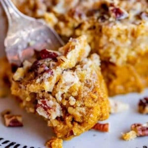 A close-up of a fork cutting into a slice of pumpkin dump cake topped with chopped pecans and crumbly streusel on a white plate with a patterned rim.