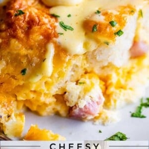 A close-up of a breakfast casserole with biscuits, ham, and eggs, topped with chopped parsley. Sauce is being drizzled over the cheesy dish. Text overlay reads: “Cheesy Ham and Egg Biscuit Breakfast Casserole Overnight Option.”.