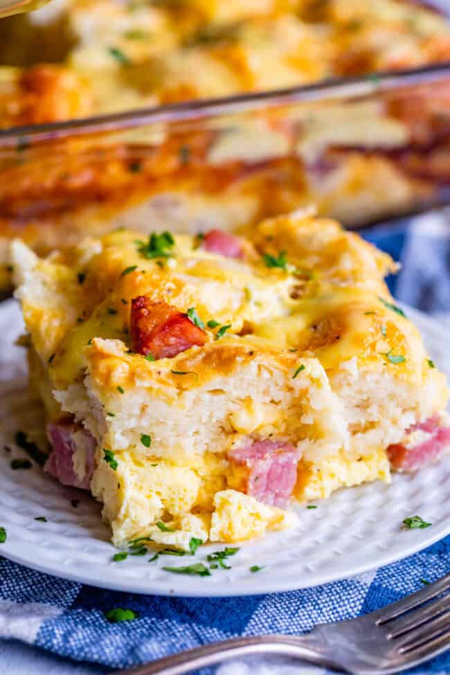 A close-up of a breakfast casserole with biscuits, featuring chunks of ham, melted cheese, and chopped herbs on a white plate. In the background, there’s a dish filled with more casserole.