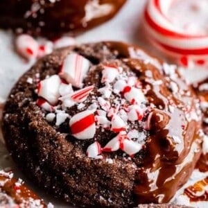 A close-up of chocolate peppermint cookies topped with melted chocolate and crushed peppermint candy, with more cookies and candy canes in the background.