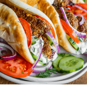 Three Greek gyros filled with savory gyro meat, sliced tomatoes, red onions, cucumbers, and tzatziki sauce on pita bread, served on a white plate with fresh herbs. Text overlay reads: Restaurant Style Greek Gyros Savory Gyro Meat.