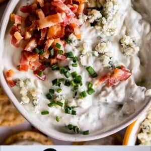 A bowl of creamy blue cheese dip topped with crumbled bacon, more blue cheese, and chopped chives, surrounded by sliced apples and crackers.