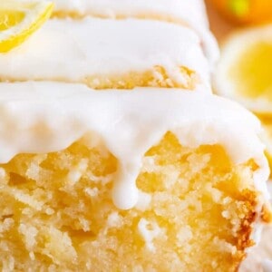 A close-up of a sliced lemon loaf cake topped with glossy white icing and garnished with lemon slices. The cake appears moist and dense with a rich, golden crumb, showcasing a classic lemon bread recipe.