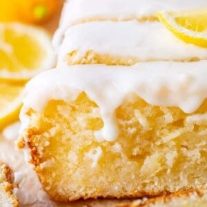 A close-up of a sliced lemon bread loaf cake topped with white icing and lemon slices, with a lemon in the background. The texture of the moist cake is visible.