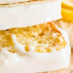 Close-up of a sliced lemon loaf cake with a thick layer of white icing on top, resting on parchment paper. Inspired by a classic lemon bread recipe, a lemon slice is visible in the background.