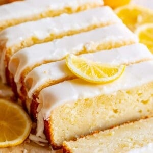 A sliced loaf of lemon bread topped with white icing and garnished with lemon slices. Whole lemons are visible in the background, perfect for fans of a classic lemon bread recipe.
