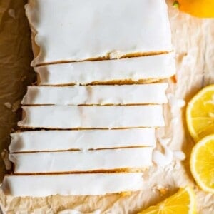 A rectangular lemon loaf cake with white glaze, sliced into pieces, sits on parchment paper with lemon slices, whole lemons, and a hint of a delicious lemon bread recipe nearby.