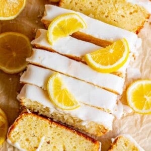 A glazed lemon bread loaf sliced on parchment paper, topped with lemon glaze and garnished with fresh lemon slices. Whole and halved lemons are scattered around the loaf.