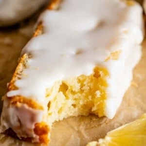 A slice of lemon cake with white icing, inspired by a classic lemon bread recipe, sits on brown parchment paper next to a few fresh lemon slices.