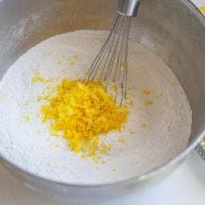 A metal mixing bowl with flour and lemon zest being whisked sits beside fresh lemons and a zester on a white surface—perfect for making your favorite lemon bread.