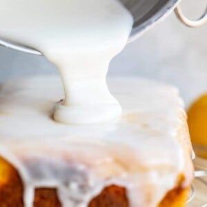 A thick white glaze is being poured from a bowl onto a rectangular lemon bread, covering the top and dripping down the sides.