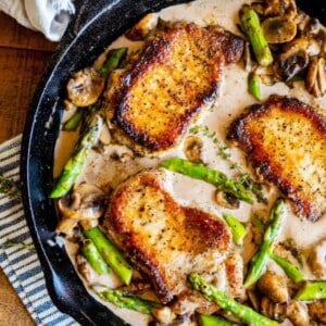 A cast iron skillet filled with pork chops in sauce, asparagus, and mushrooms sits on a striped towel on a wooden surface. A napkin and fresh thyme are nearby.