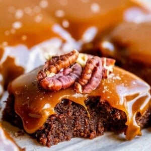 A close-up of rich salted caramel brownies topped with glossy caramel sauce and pecan halves, with more caramel and brownies blurred in the background.