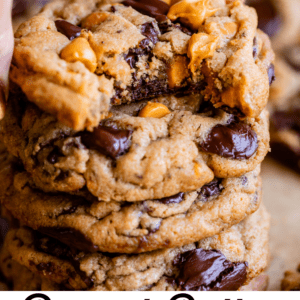 A hand lifts a thick, chewy cookie loaded with chocolate and butterscotch chips from a stack. Text below reads, Peanut Butter Butterscotch Cookies – The Food Charlatan.