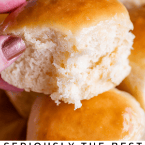 A hand holds a fluffy, golden-brown dinner roll with a bite taken out, revealing its soft interior. More rolls are piled in the background. Text reads: “One Hour Dinner Rolls—Super Soft & Fluffy! The Food Charlatan.”.