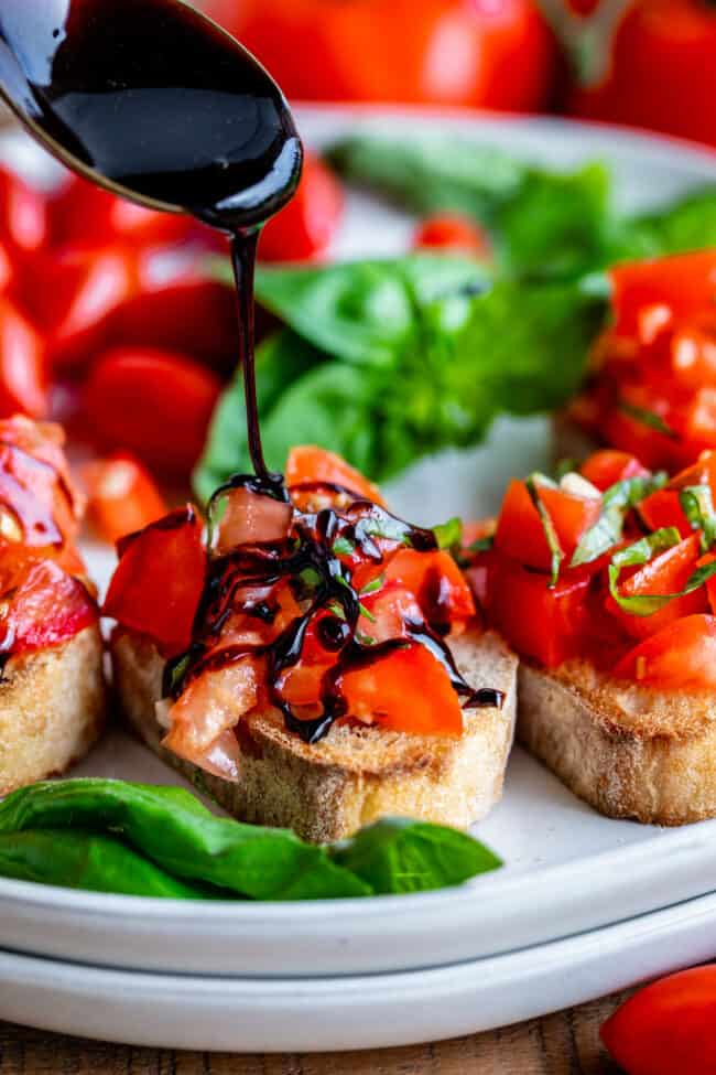 drizzling a balsamic vinegar reduction onto bruschetta on stacked white plates on a wooden cutting board.