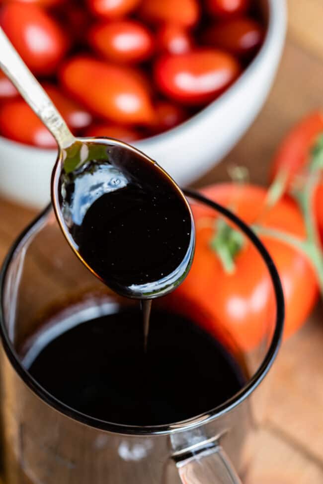 balsamic vinegar reduction drizzling from a spoon into a glass measuring cup with tomatoes in the background.