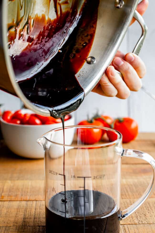 pouring a balsamic reduction from a metal pan into a glass measuring cup with tomatoes in the background. 