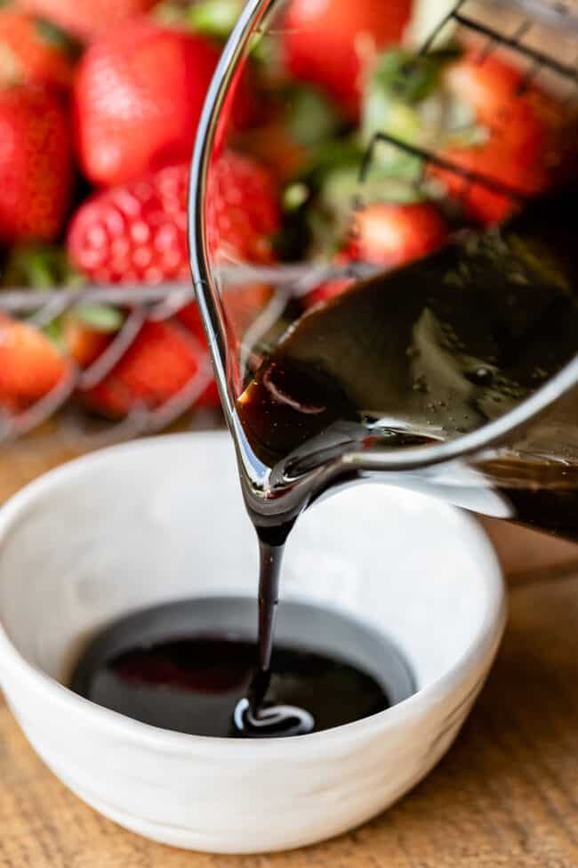 pouring balsamic vinegar reduction from glass measuring cup into white ceramic bowl with strawberries in the background.