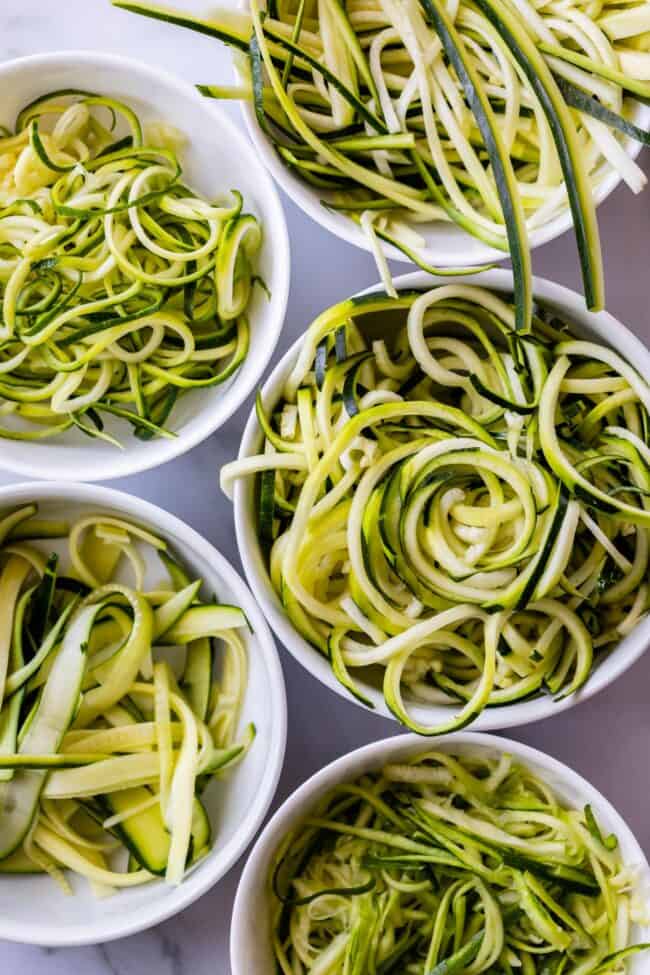 several white bowls full of zoodles made using different methods.