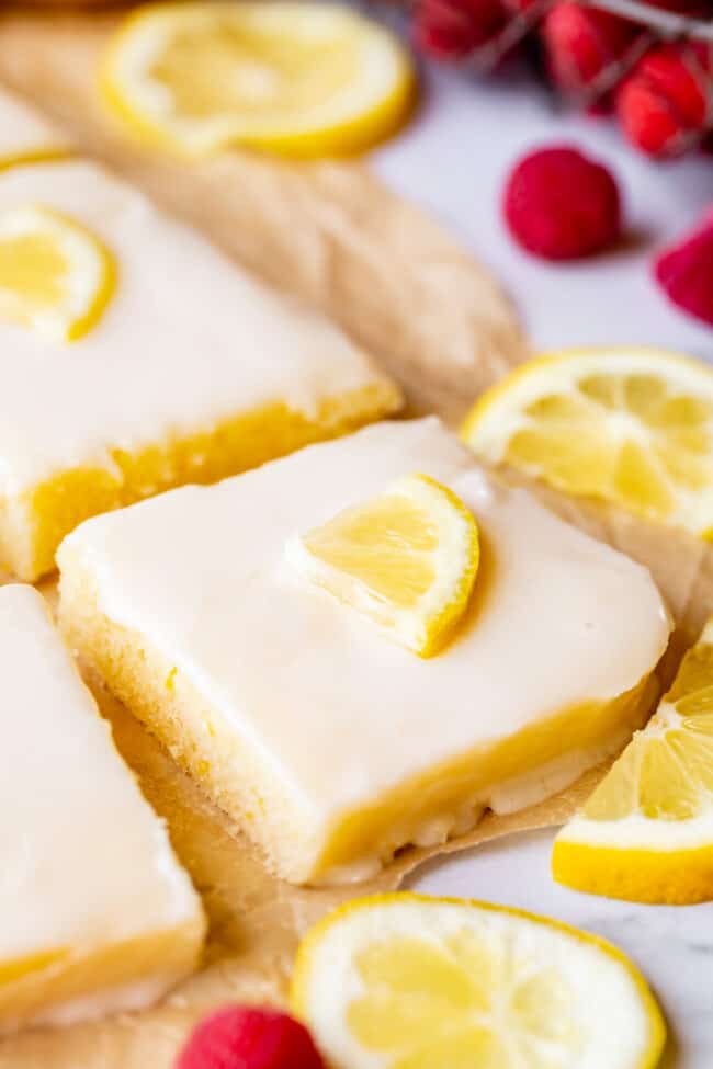 squares of lemon sheet cake with lemon slices and fresh raspberries on a parchment lined baking sheet.