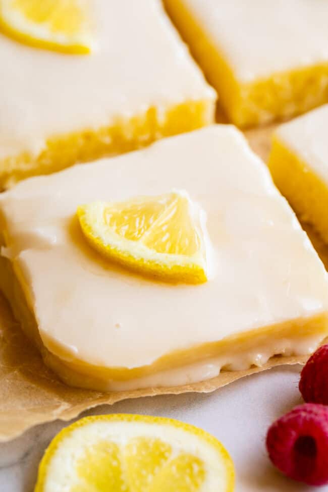 squares of lemon sheet cake with glaze, lemon slices, and fresh raspberries on parchment paper.