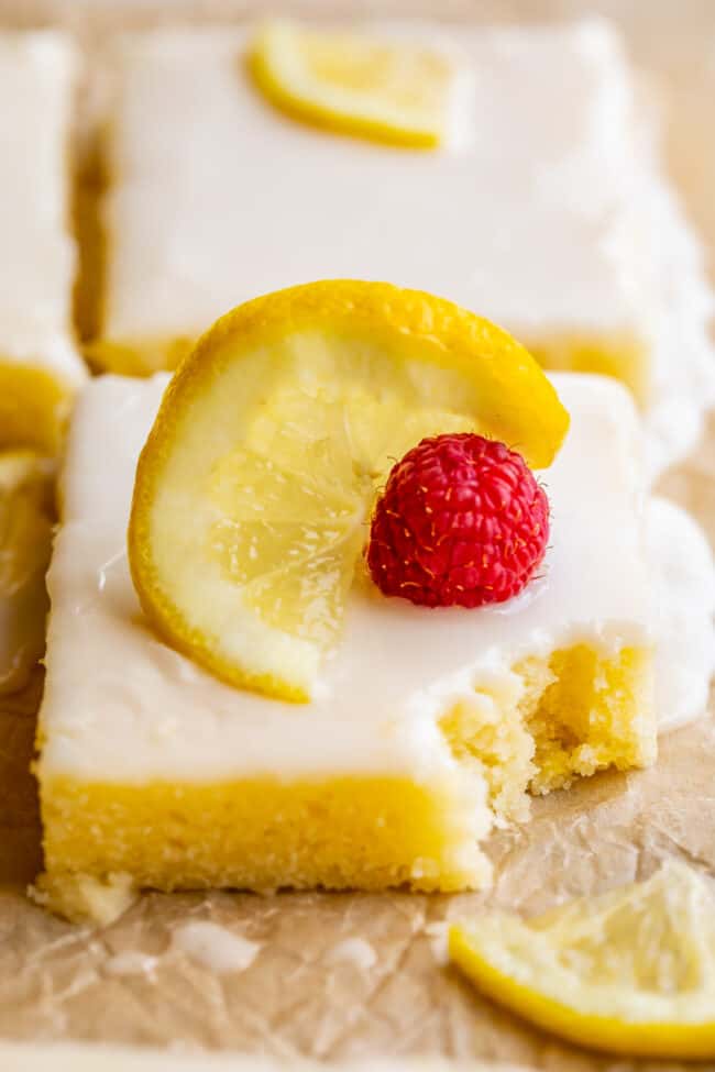 squares of lemon sheet cake with glaze, sliced lemons, and a fresh raspberry, with one bite taken from the cake