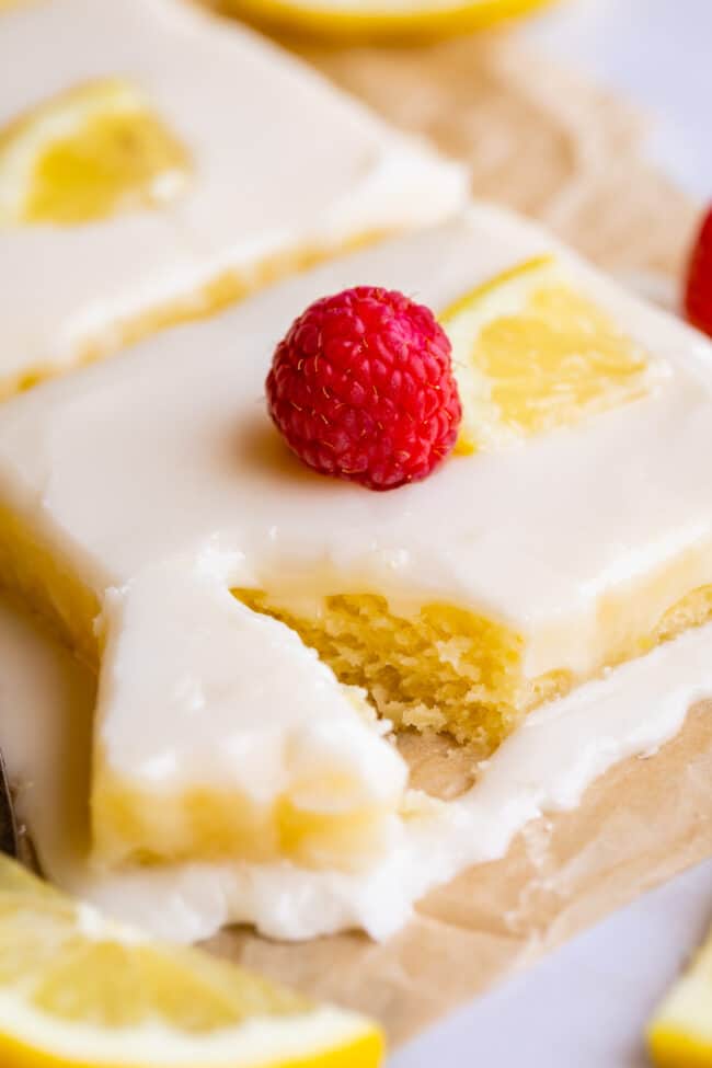 lemon sheet cake with glaze, a sliced lemon, and a fresh raspberry, one bite  partially removed.