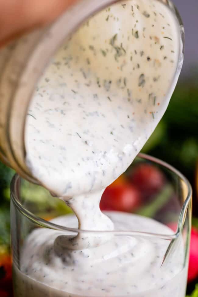 creamy buttermilk ranch recipe being poured from a glass jar into a glass measuring cup.