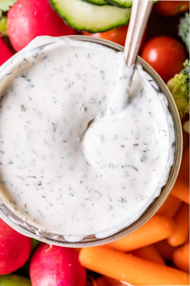 homemade buttermilk ranch dressing in glass jar with a spoon in it surrounded by vegetables. 