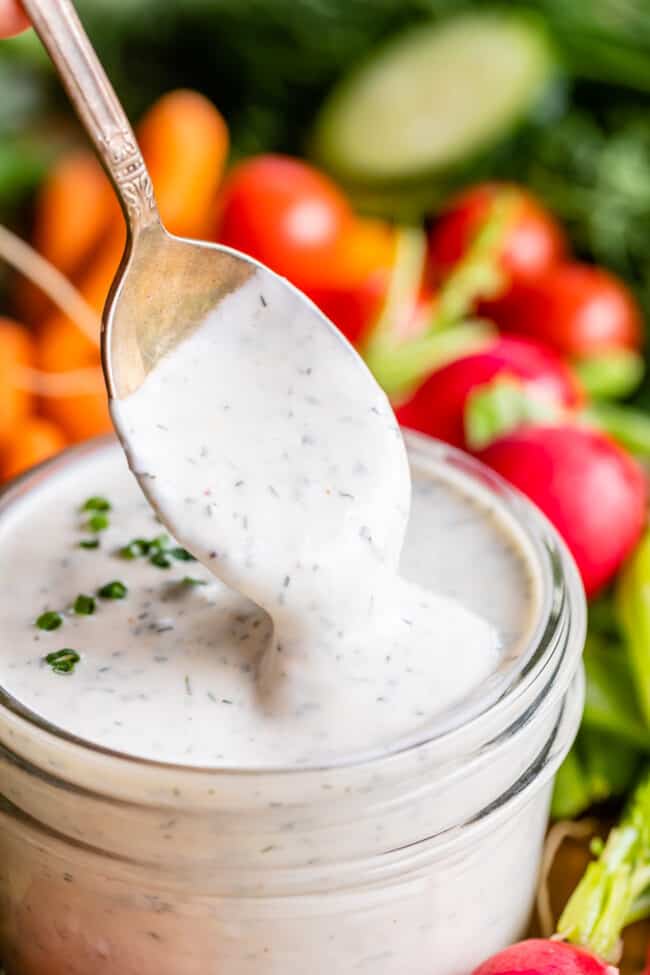 creamy buttermilk ranch dressing with spoon dipping into glass jar with fresh vegetables in the background.