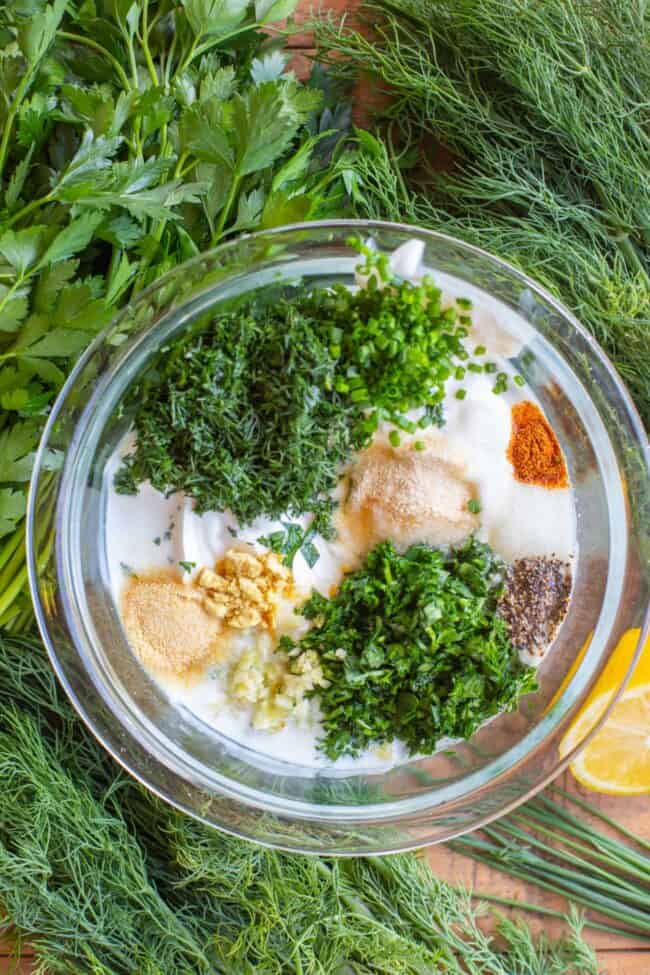 fresh herbs and spices being added to dressing in glass jar surrounded by fresh herbs and lemons. 