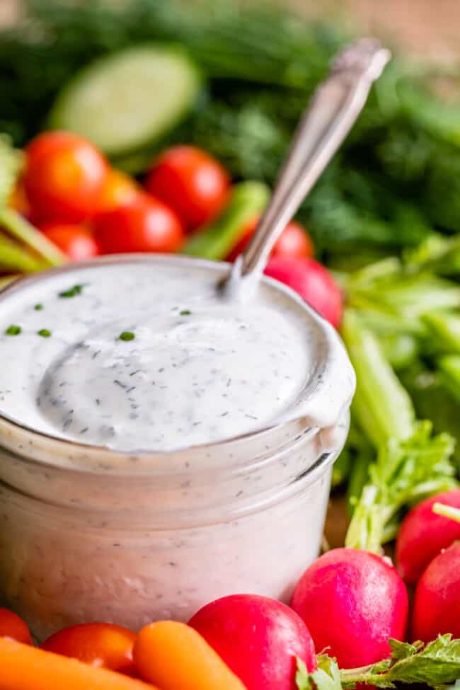 buttermilk ranch dressing in a glass jar with a spoon surrounded by fresh vegetables.