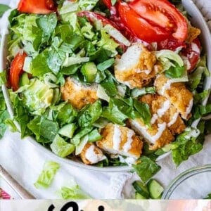 A bowl of crispy chicken salad topped with tomato wedges, lettuce, and a drizzle of buttermilk ranch dressing. A smaller bowl of sliced tomatoes and dressing sit nearby. Text reads Crispy Chicken Salad with Buttermilk Ranch.