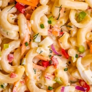 Close-up of creamy macaroni salad recipe featuring elbow pasta, chopped red onion, red bell pepper, green onions, shredded carrots, and herbs mixed in a rich mayonnaise-based dressing.