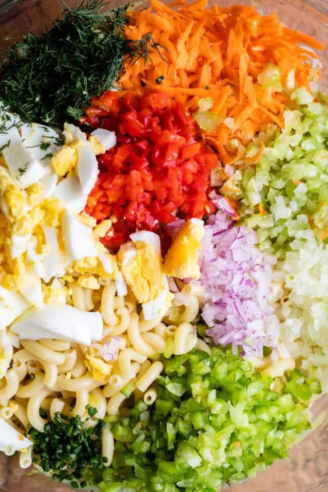 finely chopped ingredients in a bowl ready to be mixed, including hard boiled eggs, bell peppers, carrots, celery, pasta, other fresh veggies, and herbs. 