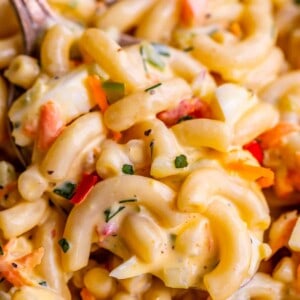 Close-up of creamy macaroni salad with elbow pasta, chopped vegetables, and herbs, all coated in a rich, yellowish dressing. A fork is partially visible, mixing this classic macaroni salad recipe.