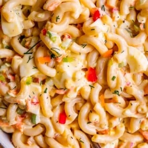 Close-up of creamy macaroni salad with elbow pasta, chopped red bell pepper, green onions, and herbs mixed in a mayonnaise-based dressing. A spoon is partially visible in the bowl, highlighting this flavorful macaroni salad.