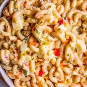 Close-up of creamy macaroni salad with elbow pasta, diced red bell peppers, chopped hard-boiled eggs, and fresh herbs mixed in a bowl, with a spoon visible in the macaroni salad.