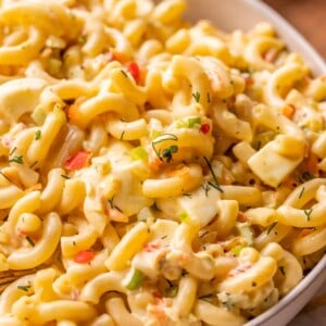 A close-up of a bowl filled with creamy macaroni salad, featuring elbow pasta, diced vegetables, herbs, and chunks of hard-boiled egg, all coated in a rich, yellowish dressing—a must-try macaroni salad recipe.