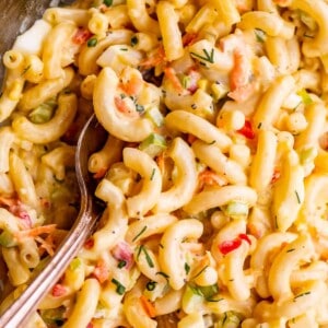 Close-up of creamy macaroni salad with elbow pasta, diced red peppers, green onions, and herbs in a rich dressing. A metal spoon rests in the bowl, ready to serve this delicious macaroni salad.