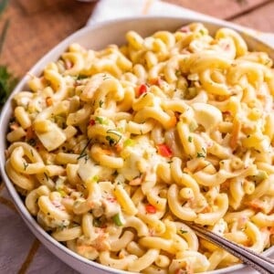 A white bowl filled with creamy macaroni salad mixed with vegetables and herbs sits on a checkered cloth with a spoon inside. A small dish of chopped onions is in the background—a perfect scene for any macaroni salad recipe.