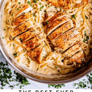 A skillet filled with creamy fettuccine Alfredo, topped with juicy, seasoned chicken breasts and garnished with chopped parsley. Text below reads: Chicken Fettuccine Alfredo - The Food Charlatan.