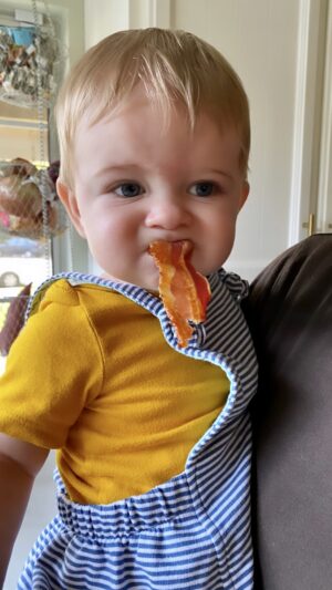 Adorable baby boy with a strip of bacon in his mouth.