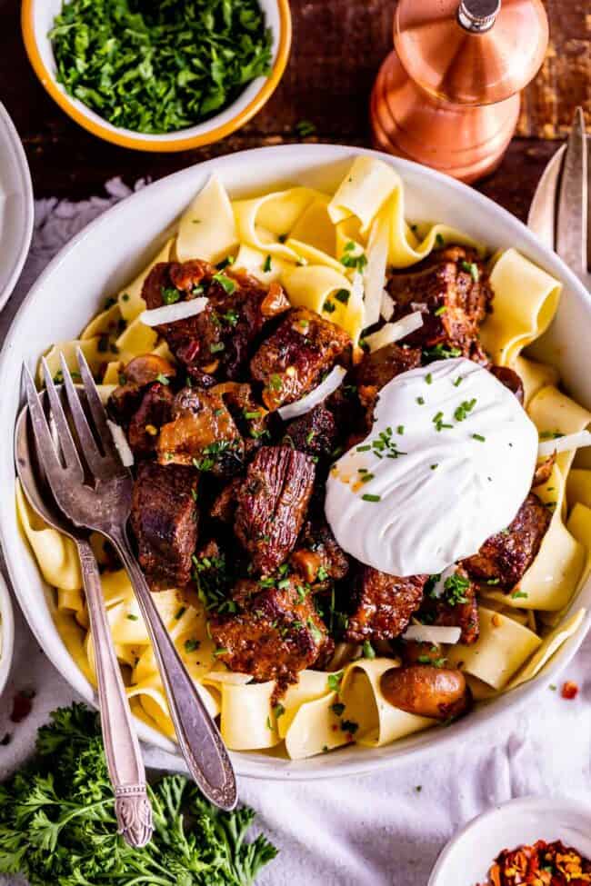 crock pot beef stroganoff with a big dollop of sour cream and fresh chives in a bowl with a fork and spoon.