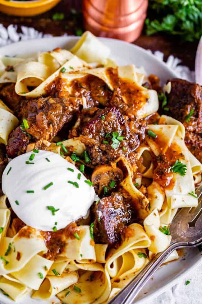 crockpot beef stroganoff served over  pappardelle pasta on a plate with sour cream and fresh chives.