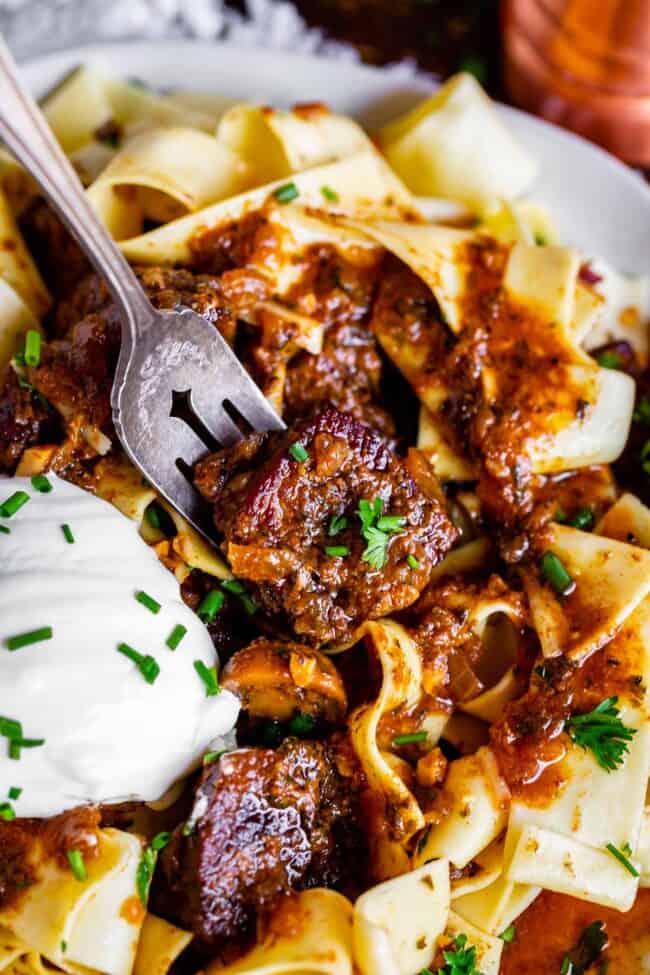 a fork lifting a bite of crock pot beef stroganoff with sour cream and fresh herbs from a plate.